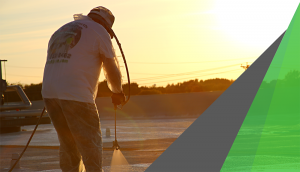 A man in a white T shirt is photographed against the sunset as he applies spray foam to a commercial roof. Spray foam works well as insulation against heat loss.